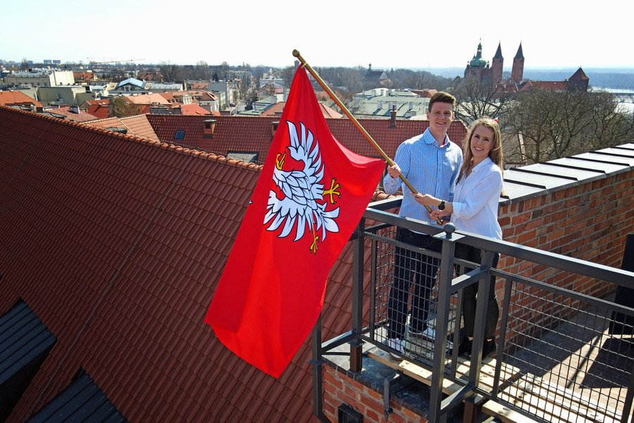 Na dachu budynku para młodych osób trzyma flagę województwa mazowieckiego