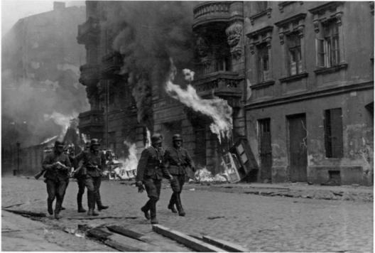 Zdjęcie z getta. Na ulicy widać kilku mężczyzn w w mundurach gestapo. W tle płonący budynek kamienicy