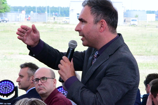 zdjęcie z profilu mężczyzny, który przemawia do grupy osób, w dłoni ma mikrofon, drugą rękę trzyma na wysokości głowy