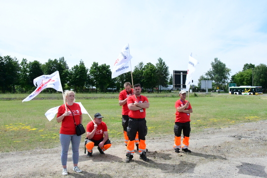 grupa osób z transparentami w dłoniach i w koszulkach z napisem SOS dla Modlina stoi na polanie.