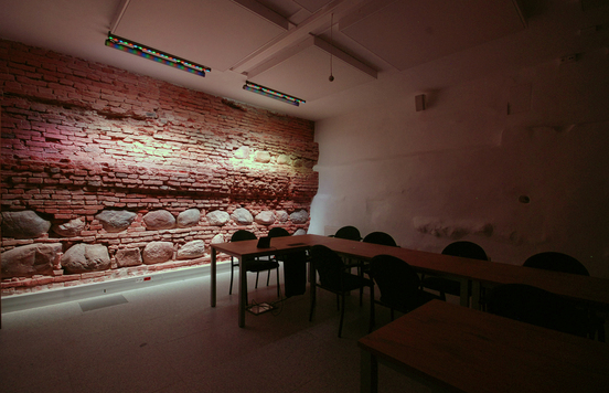 wnętrze muzeum, na pierwszym planie stoliki z krzesłami, w tle podświetlana ściana z cegieł i kamieni