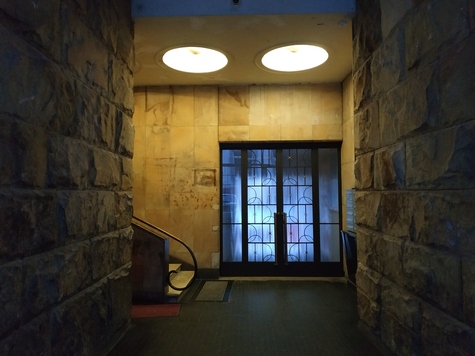 Kamienne ściany korytarza, z którego na samym końcu odchodzą w lewo schody. Na wprost są przeszkolone dwuskrzydłowe drzwi. U góry widać dwie lampy w suficie