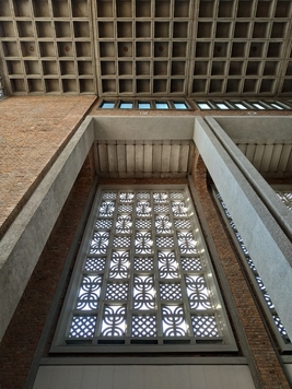 Prostokątne okno z witrażem z regularnych wzorów. Nad nim widać fragment betonowego sufitu w kwadratowe wzory, przypominające tabliczkę czekolady