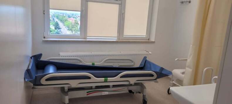 Łóżko na sali szpitalnej stoi przy oknie zasłoniętym roletami.
