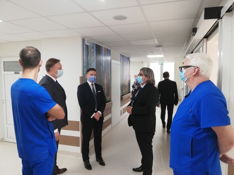 Grupa osób, wśród nich lekarze i osoby w garniturach, rozmawiają na korytarzu szpitalnym.