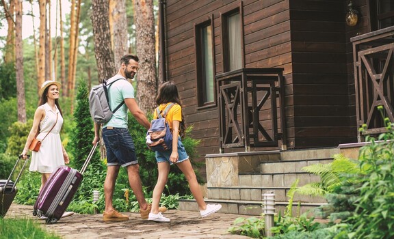 Uśmiechnięci mężczyzna i kobieta z walizkami oraz dziewczynka z plecakiem zbliżają się do schodów drewnianego domu. Zdjęcie w plenerze.