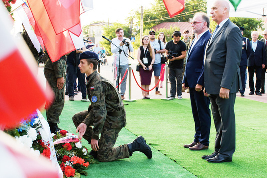 Harcerz trzymający wieniec klęczy przed pomnikiem, za nim stoi i oddaje cześć wiceprzewodniczący sejmiku z osobą towarzyszącą