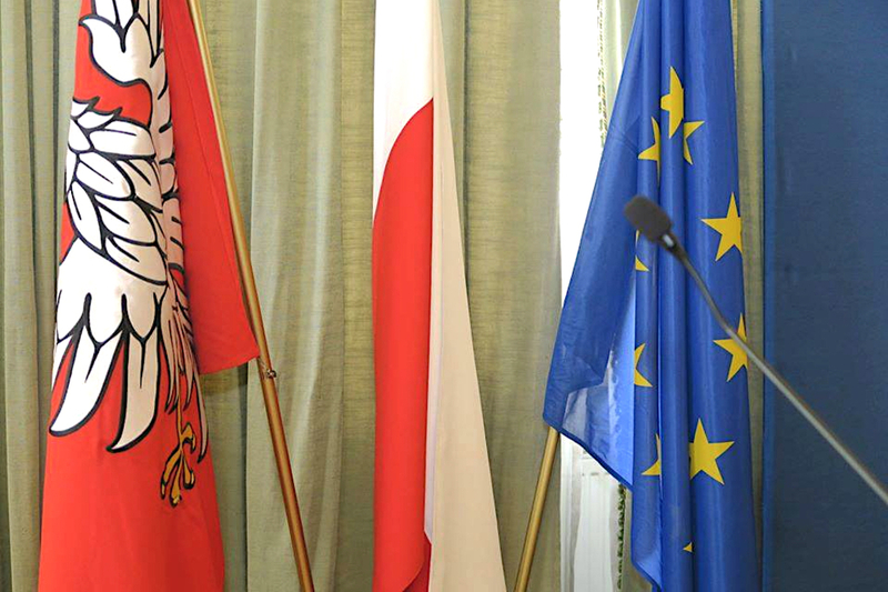Trzy flagi w stojakach na sali obrad sejmiku, na tle ściany z kotarą - od lewej: flaga Mazowsza, flaga Polski i flaga Unii Europejskiej.