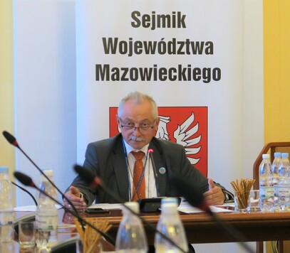 Przewodniczacy komisji siedzi za stołem obrad, za nim baner Sejmiku Województwa Mazowieckiego.
