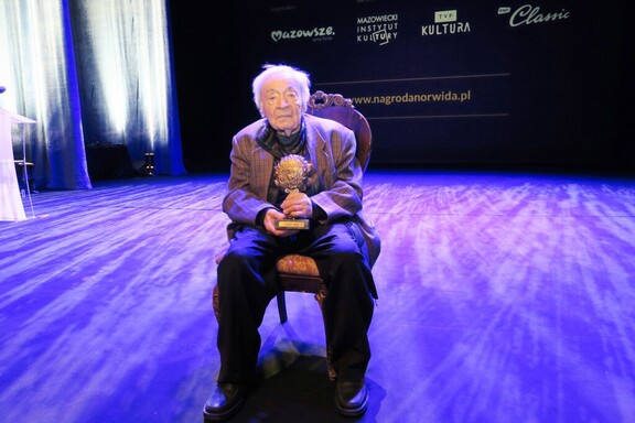 Starszy mężczyzna siedzi na krześle przodem zwrócony do kamery. W rękach trzyma statuetkę