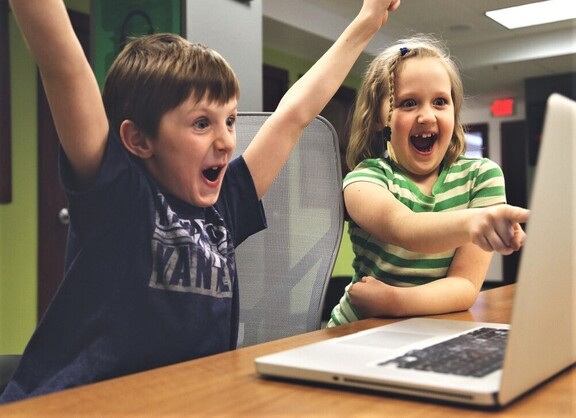 Chłopiec i dziewczynka siedzą razem przed otwartym laptopem. Dziewczynka z szeroko otwartą buzią w geście radości pokazuje palcem na monitor, chłopiec również z otwartymi ustami unosi ręce do góry w geście zwycięstwa