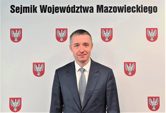 Mężczyzna w garniturze stoi na tle ścianki z napisem Sejmik Województwa Mazowieckiego i herbem Mazowsza.