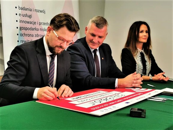 Wiceprezydent Radomia podpisuje przy stole symboliczny czek. Obok niego siedzi wicemarszałek Rajkowski, który patrzy się na niego z uśmiechem. Za wicemarszałkiem siedzi kobieta.