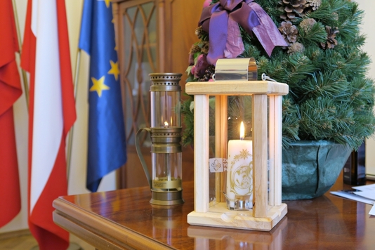 Lampion z żywym płomieniem, oraz replika lampy Ignacego Łukasiewicza – stoją na biurku, za nimi stroik choinkowy i trzy flagi: Polski, Mazowsza i Unii Europejskiej.