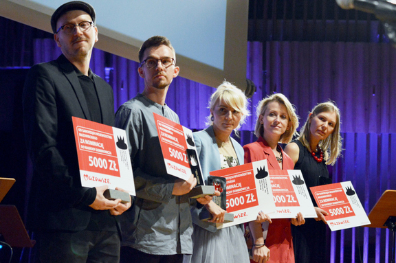Pięcioro nominowanych stoi obok siebie na scenie prezentując czeki z wygraną. Wszyscy patrzą się w bok na kamerę
