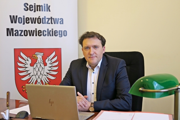 Wiceprzewodniczący sejmiku siedzi na fotelu za biurkiem przed laptopem, za nim baner z herbem Mazowsza i napisem Sejmik Województwa Mazowieckiego.