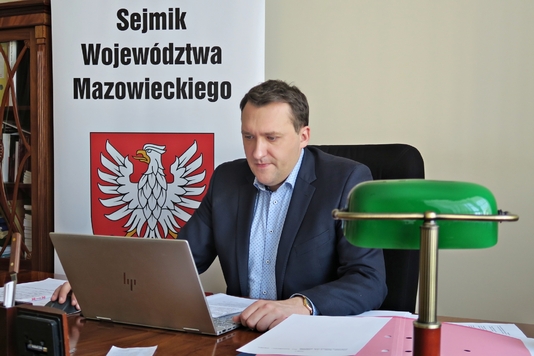 Wiceprzewodniczący siedzi za biurkiem przed laptopem, po prawej lampa biurkowa, za nim baner z herbem Mazowsza i napisem Sejmik Województwa Mazowieckiego