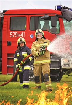 Dwójka strażaków z prądownicą na tle wozu bojowego