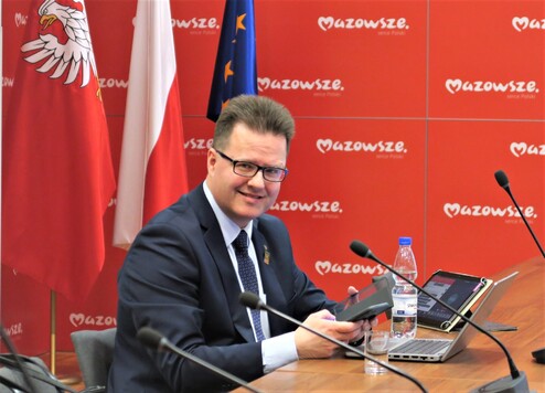 Radny siedzi za stołem przed laptopem, tabletem i mikrofonem, za nim po prawej tablica z logotypem samorządu Mazowsza, a po lewej flagi Mazowsza, Polski i Unii Europejskiej.