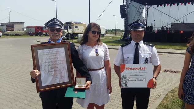 Janina Ewa Orzełowska pozuje do zdjęcia z dwoma strażakami, którzy trzymają dyplom i medal