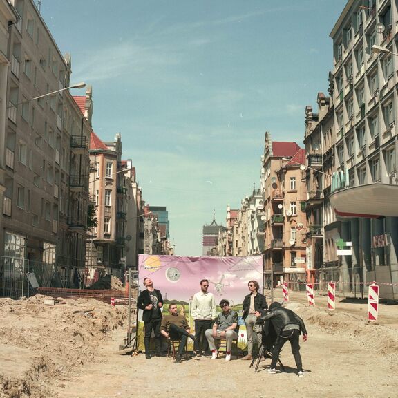 Członkowie zespołu Muchy pozują do zdjęcia na remontowanej ulicy na tle baneru z różowym niebem