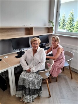 Elżbieta Lanc i jakaś kobieta siedzą bokiem przy stanowiskach z komputerami. Uśmiechają się i pozują do zdjęcia