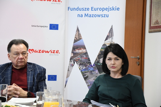 Dwie osoby siedzą przy stole, w tle ścianki z logo Mazowsze i Fundusze Europejskie na Mazowszu